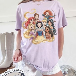 Op de afbeelding: Een lichtpaars T-shirt met een afbeelding van tekenfilmprinsessen. De kleurrijke illustratie toont verschillende Disney-prinsessen. Het T-shirt heeft een ronde hals en korte mouwen. De tekst "Comfort Colors Orchid" is op een label gedrukt.
