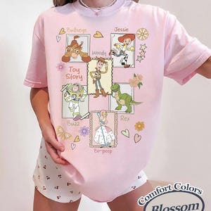 Puede incluir: Camiseta rosa claro con un dise&ntilde;o gr&aacute;fico de personajes de la pel&iacute;cula "Toy Story", incluyendo a Woody, Buzz Lightyear, Jessie, Rex, Bullseye y Bo-Peep. El dise&ntilde;o tambi&eacute;n incluye el texto "Toy Story" y "Comfort Colors Blossom".