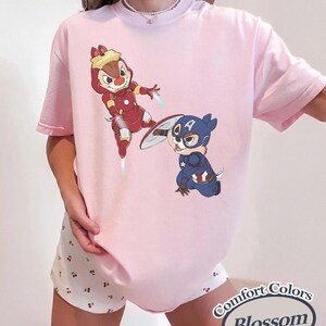 May include: A light pink t-shirt featuring cartoon characters dressed as superheroes. One character is in a red Iron Man suit, and the other is in a Captain America suit. The shirt has the words "Comfort Colors Blossom" in a circular design.