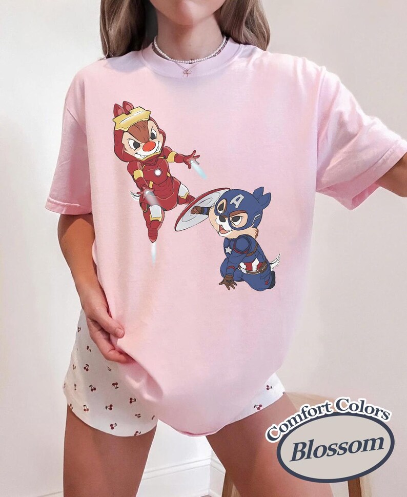 May include: A light pink t-shirt featuring cartoon characters dressed as superheroes. One character is in a red Iron Man suit, and the other is in a Captain America suit. The shirt has the words "Comfort Colors Blossom" in a circular design.