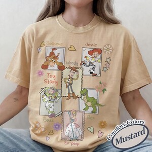 Puede incluir: Camiseta de color mostaza con un gr&aacute;fico de los personajes de Toy Story: Woody, Buzz Lightyear, Jessie, Rex, Bo-Peep y Bullseye. El dise&ntilde;o incluye detalles florales y de coraz&oacute;n, con las palabras "Toy Story" impresas.