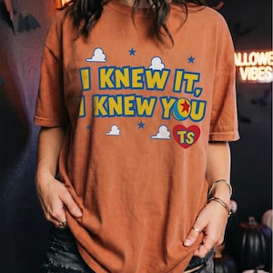 May include: A rust-colored short-sleeve t-shirt featuring the text I KNEW IT, I KNEW YOU in yellow and blue letters. The design includes small white clouds, blue stars, and a red heart with the letters TS inside, referencing a ball graphic.