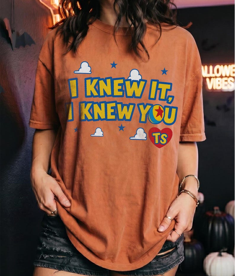 May include: A rust-colored short-sleeve t-shirt featuring the text I KNEW IT, I KNEW YOU in yellow and blue letters. The design includes small white clouds, blue stars, and a red heart with the letters TS inside, referencing a ball graphic.