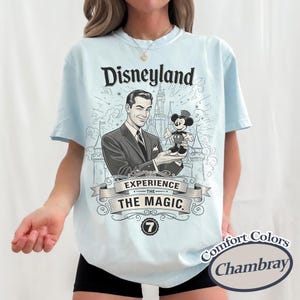 Pu&ograve; includere: T-shirt azzurra con grafica Disneyland. La grafica presenta un'illustrazione vintage di un uomo che tiene una figura di Topolino davanti a un castello, con la scritta "Disneyland Experience The Magic". La maglietta ha anche la scritta "Comfort Colors Chambray".