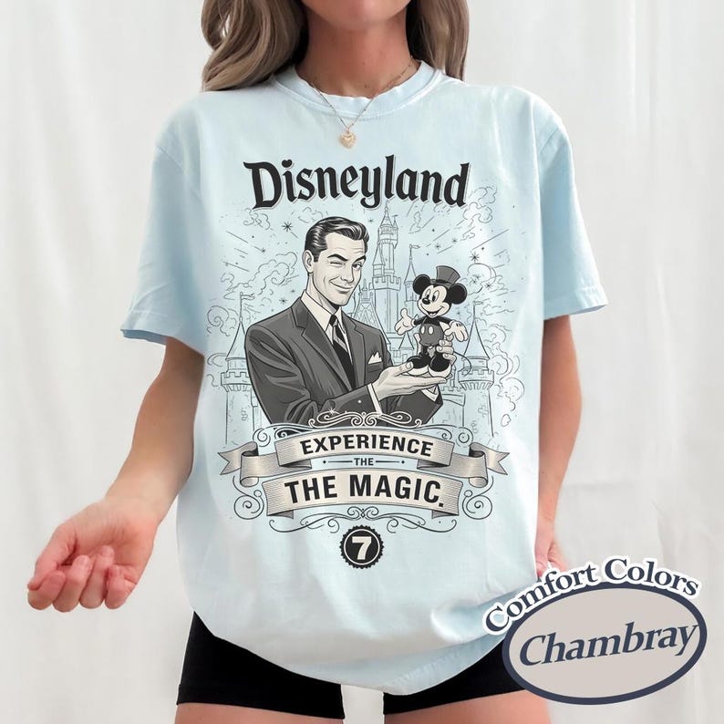 Pu&ograve; includere: T-shirt azzurra con grafica Disneyland. La grafica presenta un'illustrazione vintage di un uomo che tiene una figura di Topolino davanti a un castello, con la scritta "Disneyland Experience The Magic". La maglietta ha anche la scritta "Comfort Colors Chambray".