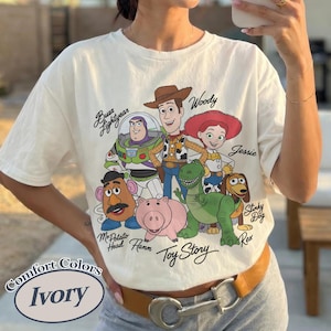Puede incluir: Camiseta de color marfil con un gr&aacute;fico de los personajes de Toy Story, incluyendo a Woody, Buzz Lightyear y Jessie. La camiseta tiene las palabras "Toy Story" y "Comfort Colors" impresas.