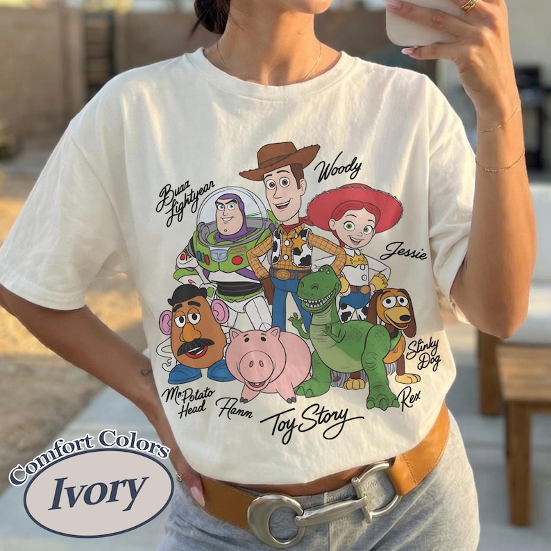 Puede incluir: Camiseta de color marfil con un gr&aacute;fico de los personajes de Toy Story, incluyendo a Woody, Buzz Lightyear y Jessie. La camiseta tiene las palabras "Toy Story" y "Comfort Colors" impresas.