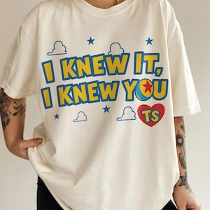 May include: An off-white short-sleeve t-shirt featuring the blue and yellow text "I KNEW IT, I KNEW YOU" with a red heart containing the letters "TS". The design includes blue stars and white cloud outlines, with a ball graphic replacing the letter O.