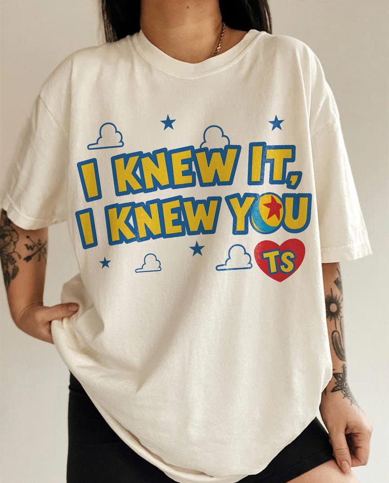 May include: An off-white short-sleeve t-shirt featuring the blue and yellow text "I KNEW IT, I KNEW YOU" with a red heart containing the letters "TS". The design includes blue stars and white cloud outlines, with a ball graphic replacing the letter O.