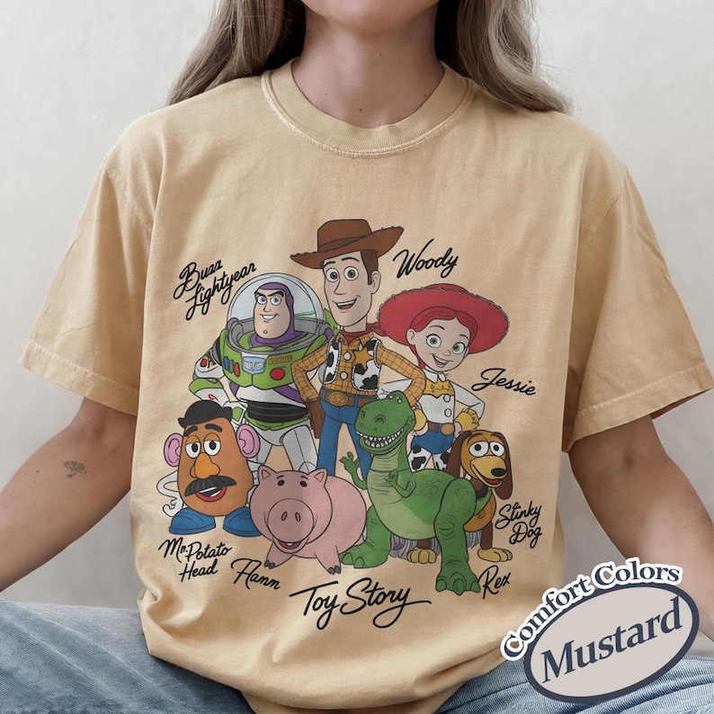 Puede incluir: Camiseta de color mostaza con un gr&aacute;fico de los personajes de Toy Story, incluyendo a Woody, Buzz Lightyear y Jessie. La camiseta tiene las palabras "Toy Story" y los nombres de los personajes impresos. La camiseta est&aacute; etiquetada como "Comfort Colors".
