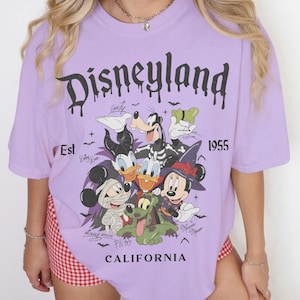 May include: A light purple t-shirt featuring a graphic print of Disney characters in Halloween costumes, including Mickey Mouse as a mummy, Minnie Mouse as a witch, and Goofy. The text reads Disneyland, Est 1955, and California in black.