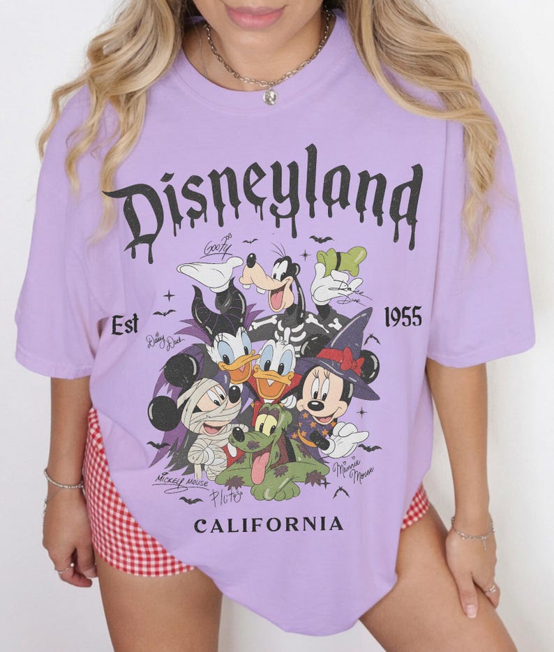 May include: A light purple t-shirt featuring a graphic print of Disney characters in Halloween costumes, including Mickey Mouse as a mummy, Minnie Mouse as a witch, and Goofy. The text reads Disneyland, Est 1955, and California in black.