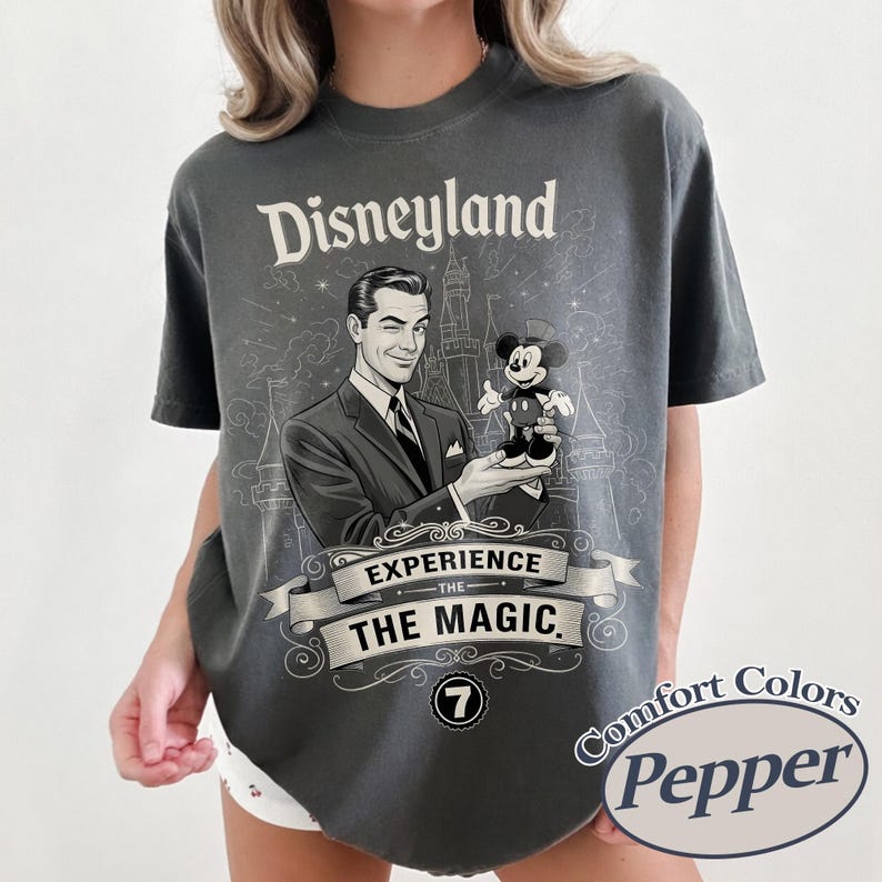 Pu&ograve; includere: T-shirt grigio scuro con grafica vintage di Disneyland. Il design include un uomo che tiene in mano Topolino, la scritta "Disneyland" e "Experience the Magic". La maglietta ha anche il logo Comfort Colors Pepper.