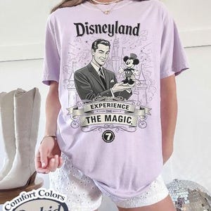 Pu&ograve; includere: Una maglietta lilla con una grafica di Disneyland. La grafica presenta un'illustrazione vintage di un uomo che tiene in mano una figura di Topolino, con le parole "Disneyland" e "Experience the Magic". La maglietta &egrave; un Comfort Colors Orchid.