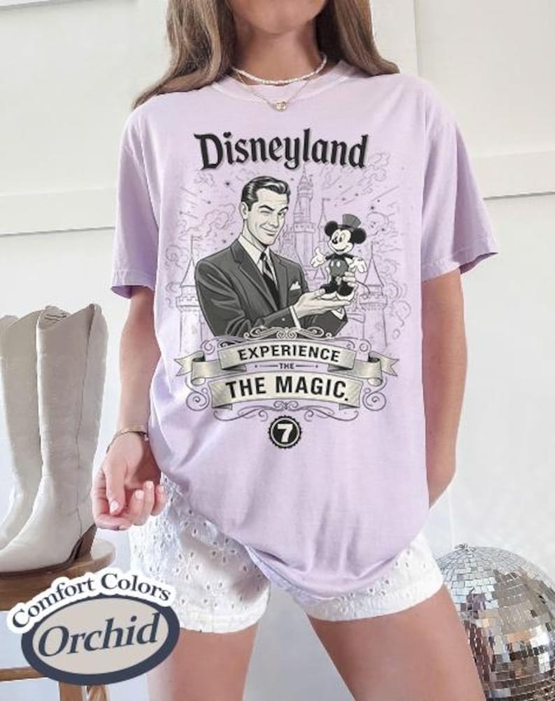Pu&ograve; includere: Una maglietta lilla con una grafica di Disneyland. La grafica presenta un'illustrazione vintage di un uomo che tiene in mano una figura di Topolino, con le parole "Disneyland" e "Experience the Magic". La maglietta &egrave; un Comfort Colors Orchid.