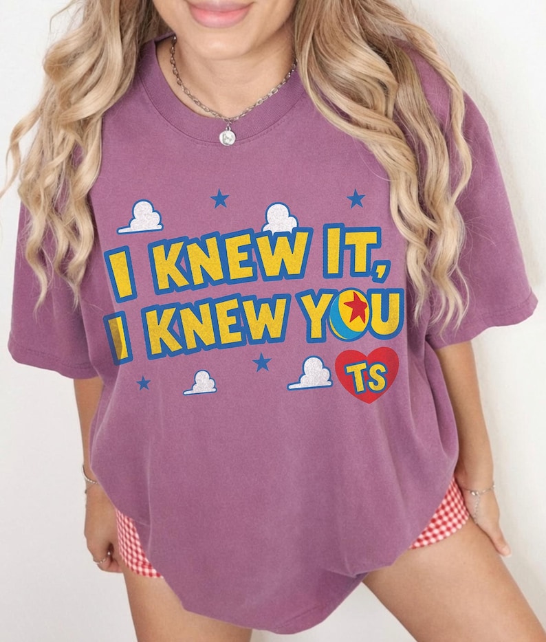 May include: A mauve short-sleeved t-shirt featuring the yellow text I KNEW IT, I KNEW YOU with a blue and yellow ball icon, a red heart with the letters TS, and small white clouds and blue stars printed across the front of the garment.