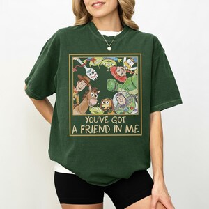 May include: Dark green t-shirt featuring a graphic of characters from the movie Toy Story. The graphic is in a gold-colored frame and includes the text "YOU'VE GOT A FRIEND IN ME". The shirt has short sleeves and a crew neck.