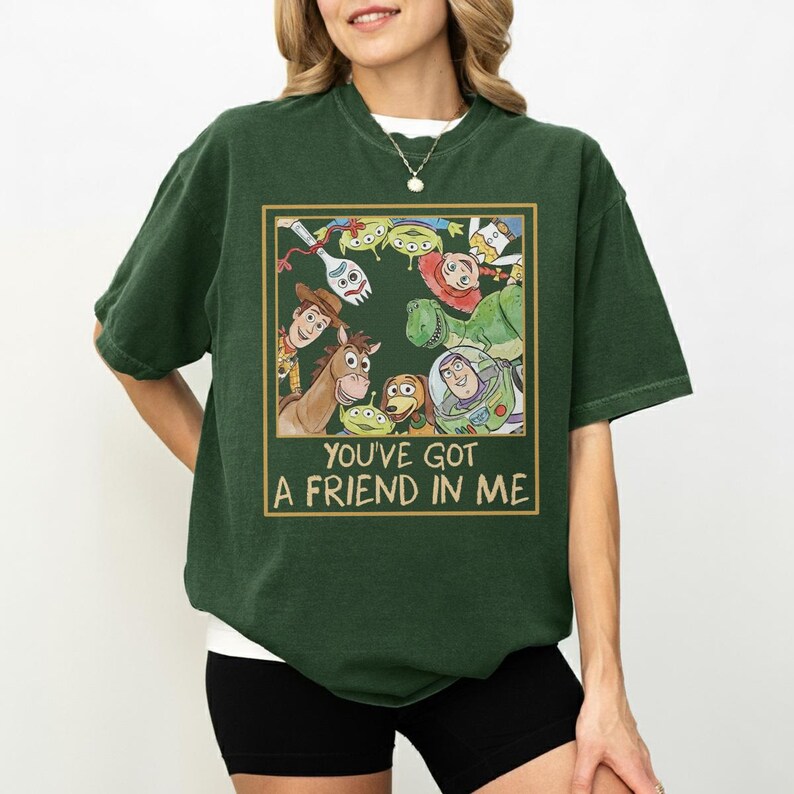 May include: Dark green t-shirt featuring a graphic of characters from the movie Toy Story. The graphic is in a gold-colored frame and includes the text "YOU'VE GOT A FRIEND IN ME". The shirt has short sleeves and a crew neck.