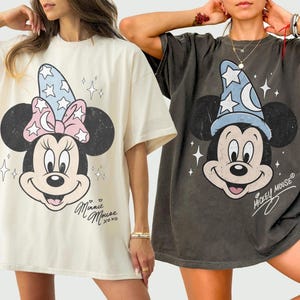May include: Two oversized t-shirts, one cream and one charcoal grey. The cream shirt features Minnie Mouse wearing a blue star-patterned hat with a pink bow and the text Minnie Mouse xoxo. The grey shirt shows Mickey Mouse in a blue wizard hat.