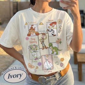 Puede incluir: Camiseta color marfil con un gr&aacute;fico de personajes de Toy Story: Bullseye, Jessie, Woody, Buzz, Rex y Bo-peep. La camiseta tiene las palabras "Toy Story" y "Comfort Colors Ivory" impresas. Se ve un cintur&oacute;n marr&oacute;n con hebilla plateada.