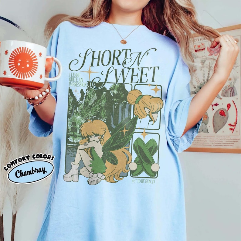 Puede incluir: Camiseta azul claro con el texto "SHORT N SWEET" y un gr&aacute;fico de un hada con alas verdes y una escena de cascada. La camiseta tambi&eacute;n tiene el texto "(6" TO BE EXACT)" y "I LEAVE QUITE AN IMPRESSION".