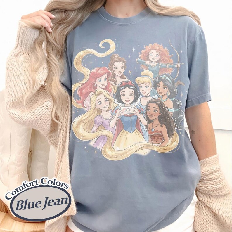 Op de afbeelding: Blauwe spijkerbroek Comfort Colors T-shirt met een groep cartoonprinsessen. De prinsessen worden afgebeeld met lang haar en verschillende outfits. Het shirt heeft een ronde hals en korte mouwen.