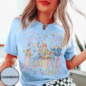 以下が含まれることがあります： 水色のTシャツ。映画「ズートピア」のキャラクター、お城、そして「Birthday Girl」の文字がカラフルに描かれています。デザインには名前と数字の「4」を入れられるスペースがあります。Tシャツは丸首で半袖です。