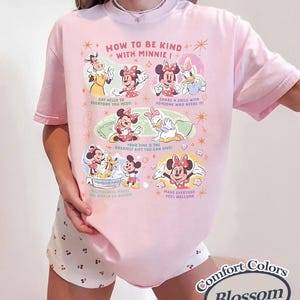 Puede incluir: Camiseta rosa claro con el texto "HOW TO BE KIND WITH MINNIE!" e ilustraciones de Minnie Mouse y sus amigos. La camiseta tambi&eacute;n muestra las palabras "Comfort Colors" y "Blossom".