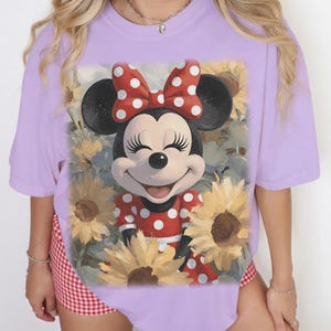 Puede incluir: Una camiseta de manga corta de color morado claro con un estampado gr&aacute;fico de Minnie Mouse con un vestido y un lazo de lunares rojos y blancos, rodeada de girasoles amarillos. El personaje tiene orejas negras y una expresi&oacute;n sonriente.