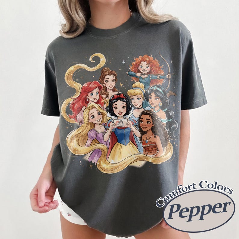 Op de afbeelding: Donkergrijs T-shirt met een cartoon illustratie van prinsessen, waaronder Sneeuwwitje en Ariel. Het shirt heeft de woorden "Comfort Colors Pepper" in een cirkelvormig ontwerp.