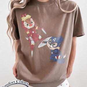 May include: Espresso-colored t-shirt featuring cartoon characters dressed as superheroes. One character is in a red and gold Iron Man suit, and the other is in a blue and white Captain America suit. The shirt has the words "Comfort Colors Espresso" printed on it.