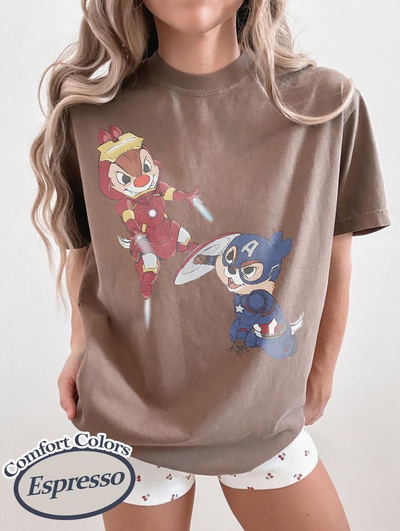May include: Espresso-colored t-shirt featuring cartoon characters dressed as superheroes. One character is in a red and gold Iron Man suit, and the other is in a blue and white Captain America suit. The shirt has the words "Comfort Colors Espresso" printed on it.