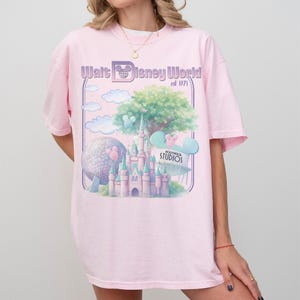 Puede incluir: Camiseta rosa claro con un gr&aacute;fico del castillo de Cenicienta, un &aacute;rbol grande y una esfera. El texto "Walt Disney World est 1971" est&aacute; impreso encima del gr&aacute;fico. La camiseta tiene mangas cortas.