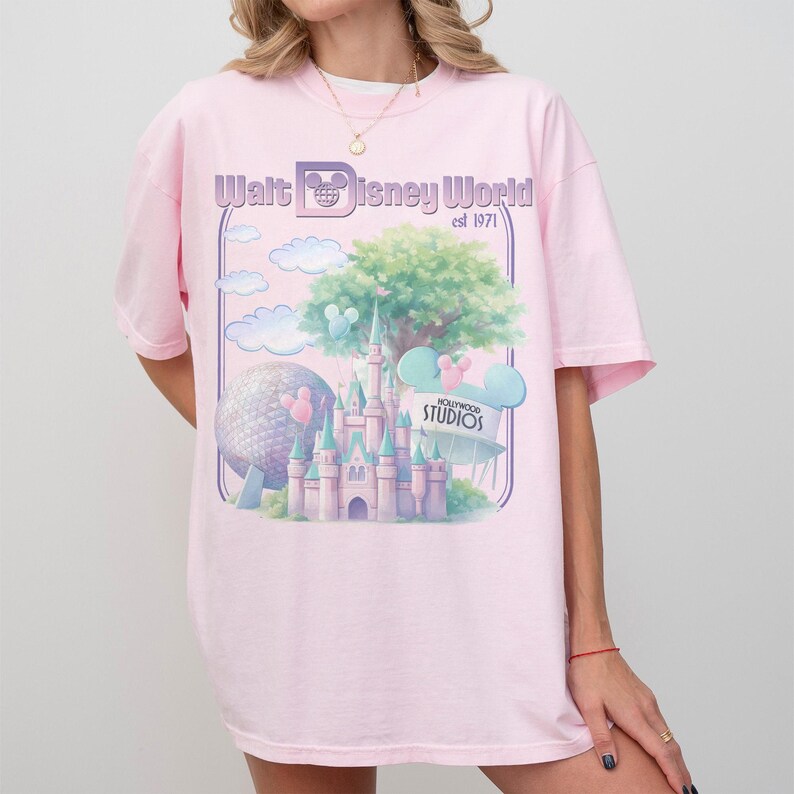 Puede incluir: Camiseta rosa claro con un gr&aacute;fico del castillo de Cenicienta, un &aacute;rbol grande y una esfera. El texto "Walt Disney World est 1971" est&aacute; impreso encima del gr&aacute;fico. La camiseta tiene mangas cortas.