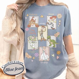 Puede incluir: Camiseta azul vaquero Comfort Colors con ilustraciones de los personajes de Toy Story: Woody, Buzz, Jessie, Rex, Bo-Peep y Bullseye. La camiseta incluye el texto "Toy Story" y los nombres de los personajes.
