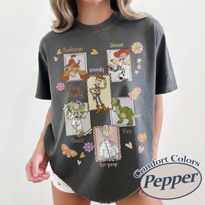 Puede incluir: Una camiseta gris oscuro con un dise&ntilde;o de personajes de Toy Story, incluyendo a Woody, Buzz Lightyear y Jessie, en un dise&ntilde;o de sello postal. La camiseta tiene las palabras "Toy Story" y "Comfort Colors Pepper" impresas.
