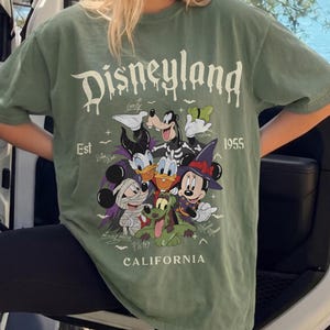 May include: A sage green short-sleeve t-shirt featuring a graphic print of Disney characters in Halloween costumes. The text reads Disneyland, Est 1955, and California, accompanied by signatures of Goofy, Daisy Duck, Mickey Mouse, Pluto, and Minnie Mouse.