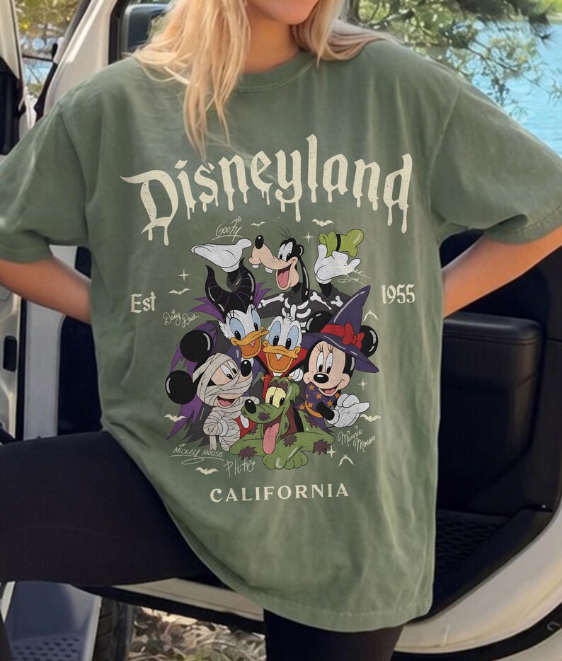 May include: A sage green short-sleeve t-shirt featuring a graphic print of Disney characters in Halloween costumes. The text reads Disneyland, Est 1955, and California, accompanied by signatures of Goofy, Daisy Duck, Mickey Mouse, Pluto, and Minnie Mouse.