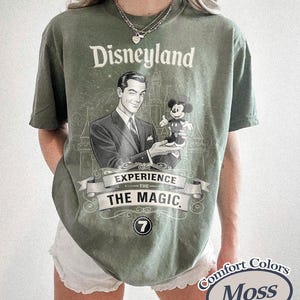 Pu&ograve; includere: T-shirt verde muschio con grafica vintage di Disneyland. Il design presenta un uomo che tiene Topolino, con le parole "Disneyland" e "Experience the Magic". La maglietta ha un girocollo e maniche corte.