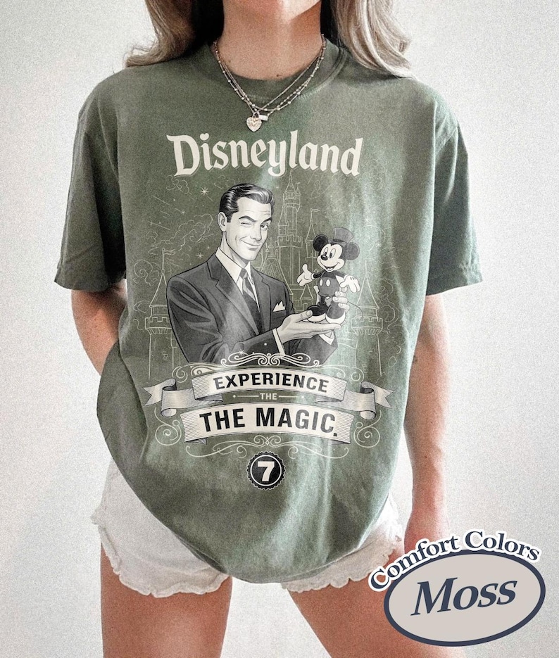 Pu&ograve; includere: T-shirt verde muschio con grafica vintage di Disneyland. Il design presenta un uomo che tiene Topolino, con le parole "Disneyland" e "Experience the Magic". La maglietta ha un girocollo e maniche corte.