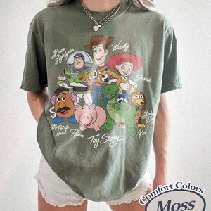 Puede incluir: Camiseta verde musgo con un gr&aacute;fico de personajes de la pel&iacute;cula animada "Toy Story". El dise&ntilde;o incluye a Woody, Buzz Lightyear, Jessie y otros personajes. La camiseta est&aacute; etiquetada como "Comfort Colors Moss".