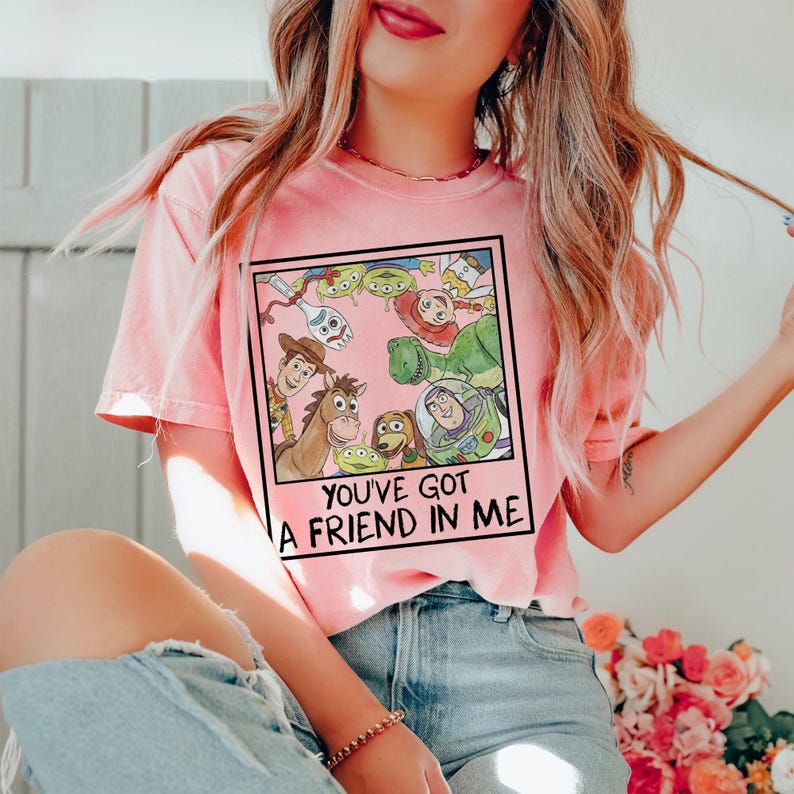 May include: Pink t-shirt featuring a graphic of characters from the animated film Toy Story. The graphic includes Woody, Buzz Lightyear, and other characters, with the text "YOU'VE GOT A FRIEND IN ME" in a black frame.