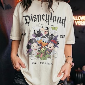 May include: A cream-colored short-sleeve t-shirt featuring a graphic of Disney characters in Halloween costumes. The text reads Disneyland Est 1955 California. The characters include Mickey Mouse as a mummy, Minnie Mouse as a witch, and Goofy.