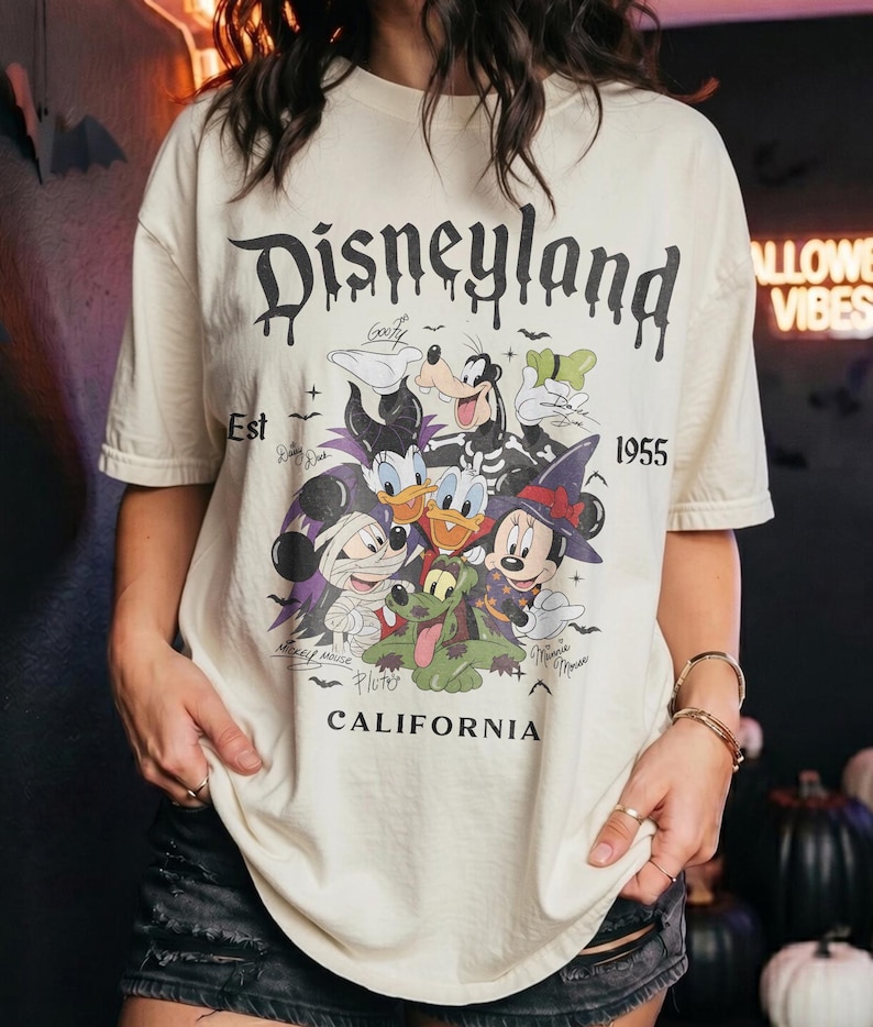 May include: A cream-colored short-sleeve t-shirt featuring a graphic of Disney characters in Halloween costumes. The text reads Disneyland Est 1955 California. The characters include Mickey Mouse as a mummy, Minnie Mouse as a witch, and Goofy.
