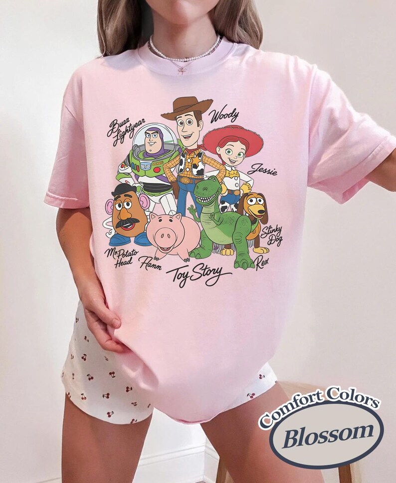 Puede incluir: Una camiseta rosa claro con un gr&aacute;fico de personajes de la pel&iacute;cula animada "Toy Story". El dise&ntilde;o incluye a Woody, Buzz Lightyear, Jessie y otros personajes. El texto "Toy Story" tambi&eacute;n es visible. La camiseta tiene mangas cortas.