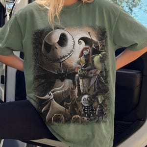 May include: A sage green short-sleeve t-shirt featuring a large, detailed graphic print of characters from The Nightmare Before Christmas, including Jack Skellington, Sally, Zero, and the trio of Lock, Shock, and Barrel against a dark background.
