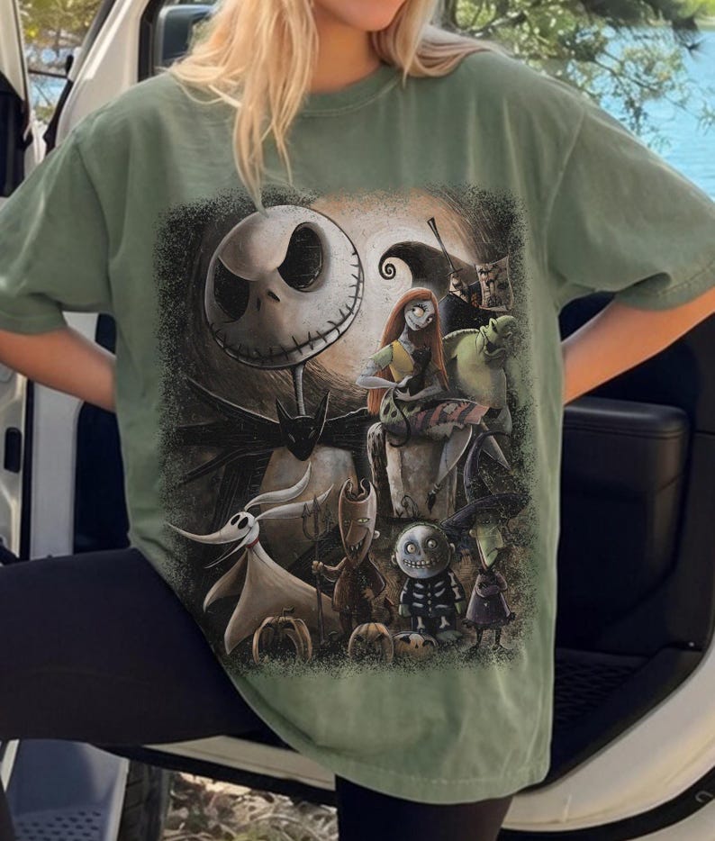 May include: A sage green short-sleeve t-shirt featuring a large, detailed graphic print of characters from The Nightmare Before Christmas, including Jack Skellington, Sally, Zero, and the trio of Lock, Shock, and Barrel against a dark background.