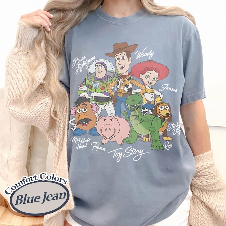 Puede incluir: Camiseta azul vaquero Comfort Colors con un gr&aacute;fico de personajes de Toy Story, incluyendo a Woody, Buzz Lightyear, Jessie y Rex. La camiseta tiene cuello redondo y mangas cortas. El texto "Toy Story" est&aacute; impreso debajo.