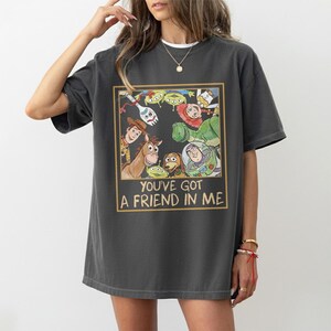 May include: Dark gray t-shirt featuring a colorful graphic of characters from the animated film Toy Story. The graphic is framed in a gold border and includes the text "YOU'VE GOT A FRIEND IN ME". The shirt has short sleeves and a relaxed fit.