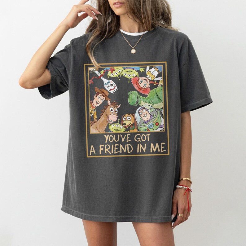 May include: Dark gray t-shirt featuring a colorful graphic of characters from the animated film Toy Story. The graphic is framed in a gold border and includes the text "YOU'VE GOT A FRIEND IN ME". The shirt has short sleeves and a relaxed fit.